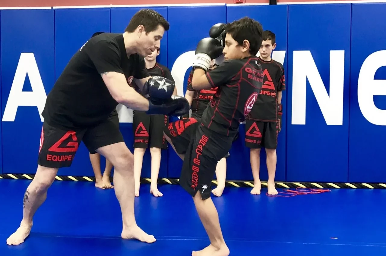 A martial arts instructor spars with a young woman practicing Muay Thai in a gym, while others watch in the background.