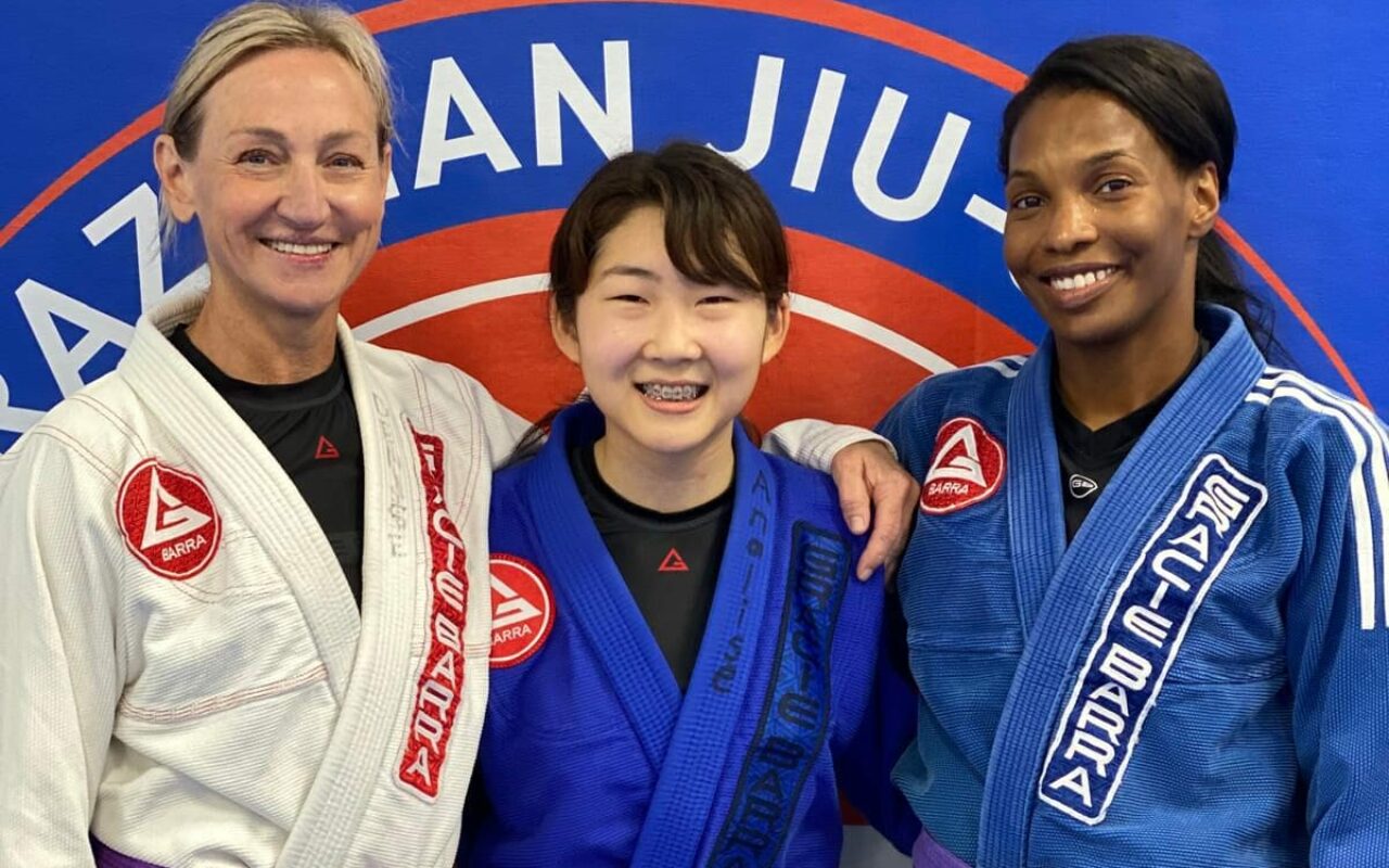 Three people in Brazilian Jiu-Jitsu uniforms stand smiling in front of a blue wall with a circular Gracie Barra logo. Two wear white gi, and one wears blue. All proudly display their purple belts.