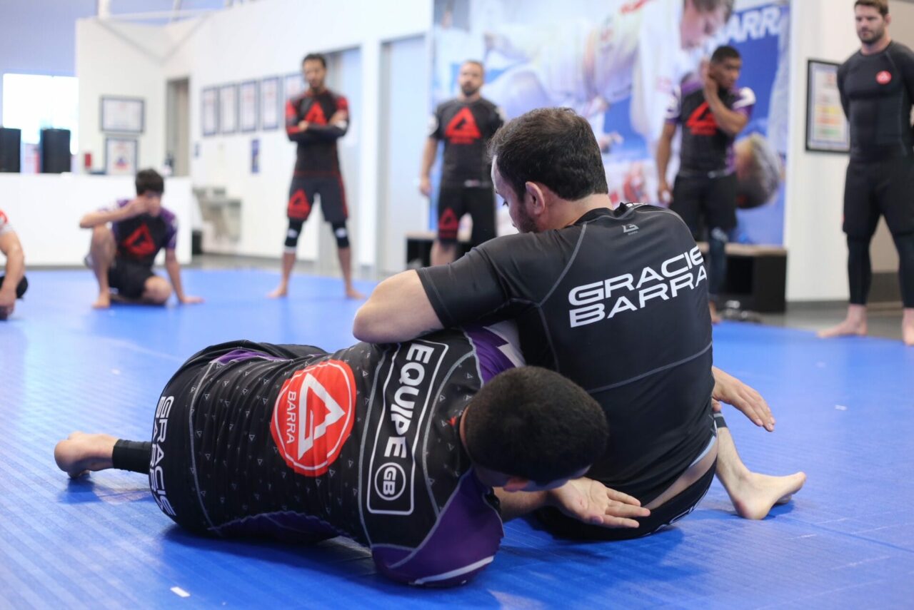 Two people in martial arts uniforms are practicing grappling on a blue mat in a gym as part of the GB¹ Program, with others observing enthusiastically in the background.