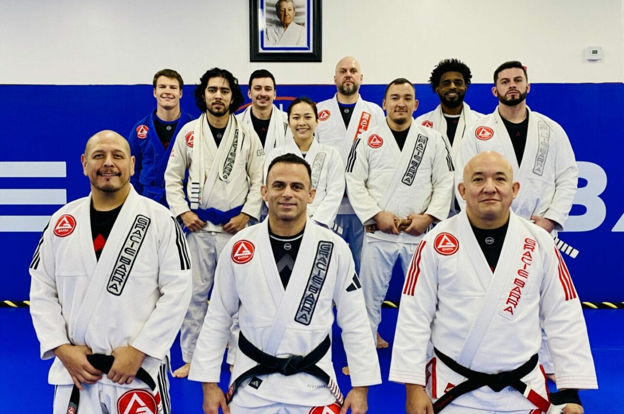 A group of martial artists, including skilled women kickboxers, pose in their white gis on a blue mat, with a framed photo on the wall behind them.