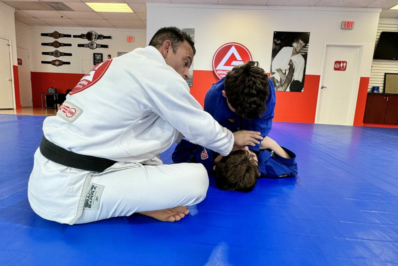 Two people in martial arts uniforms practice a technique on a blue mat in a training room with wall decorations, as part of an exclusive Private Training Program.