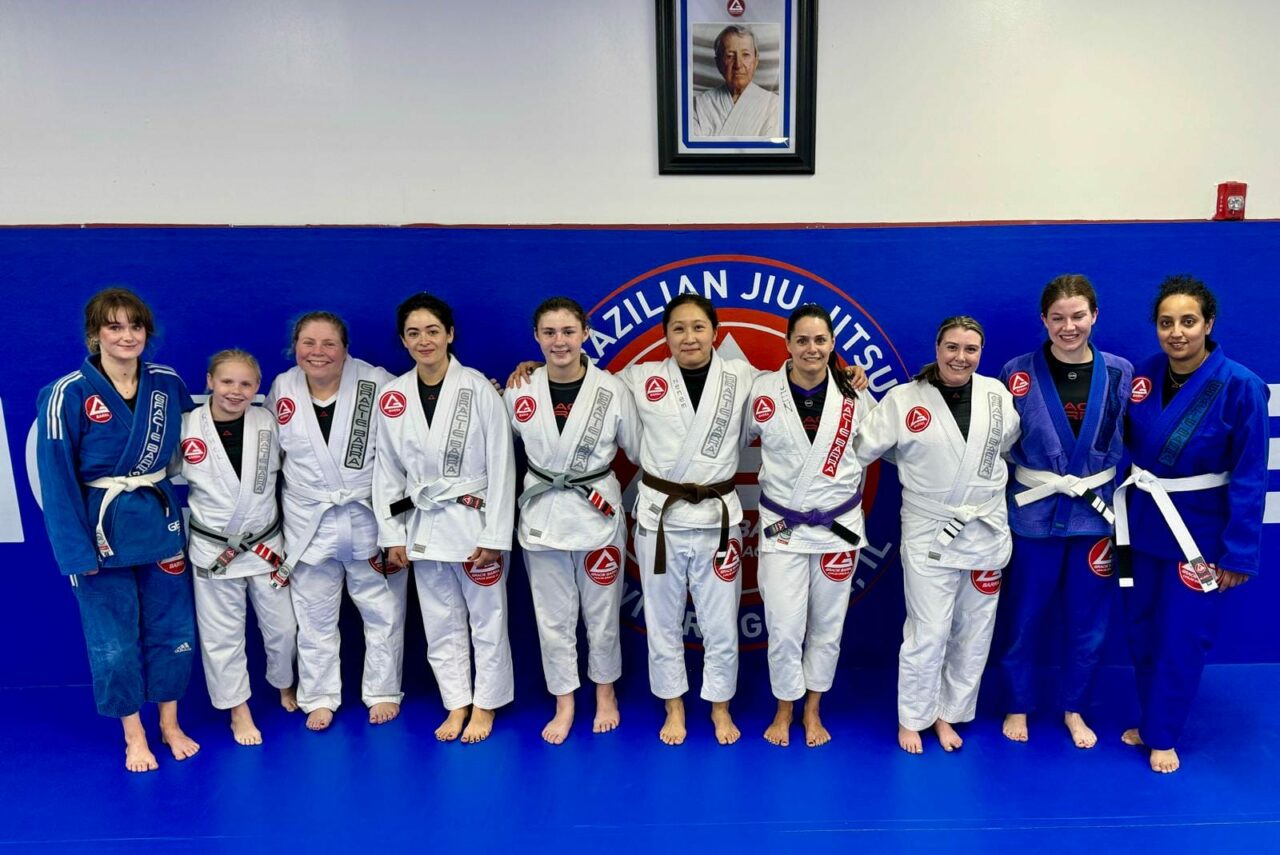 A group of people in martial arts gis stands in a dojo, under a framed photo. Some wear white gis, others wear blue, showing their dedication to the GBF Program.