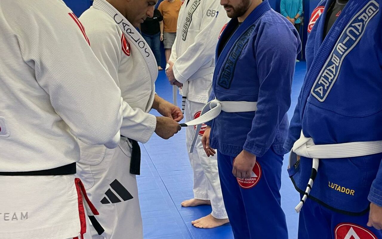 During a GB¹ Program ceremony, a martial arts instructor awards a belt to a student. Other students and instructors await in the background on a blue mat, showcasing discipline and respect.