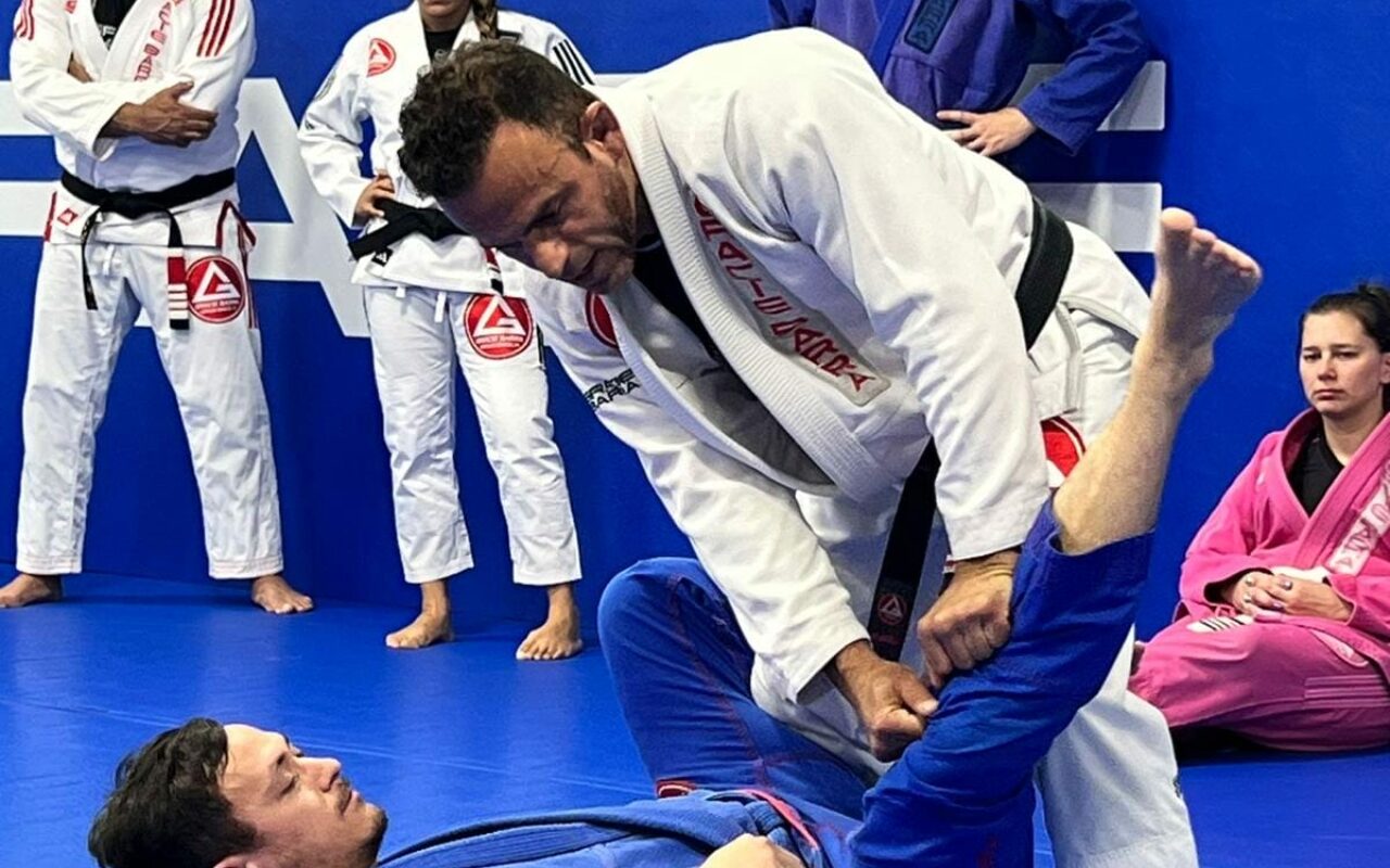 Two people engage in Brazilian Jiu-Jitsu on a blue mat, showcasing techniques from the GB³ Program, while others observe. The standing participant grips the uniform pants of the person lying on their back, illustrating control and strategy.