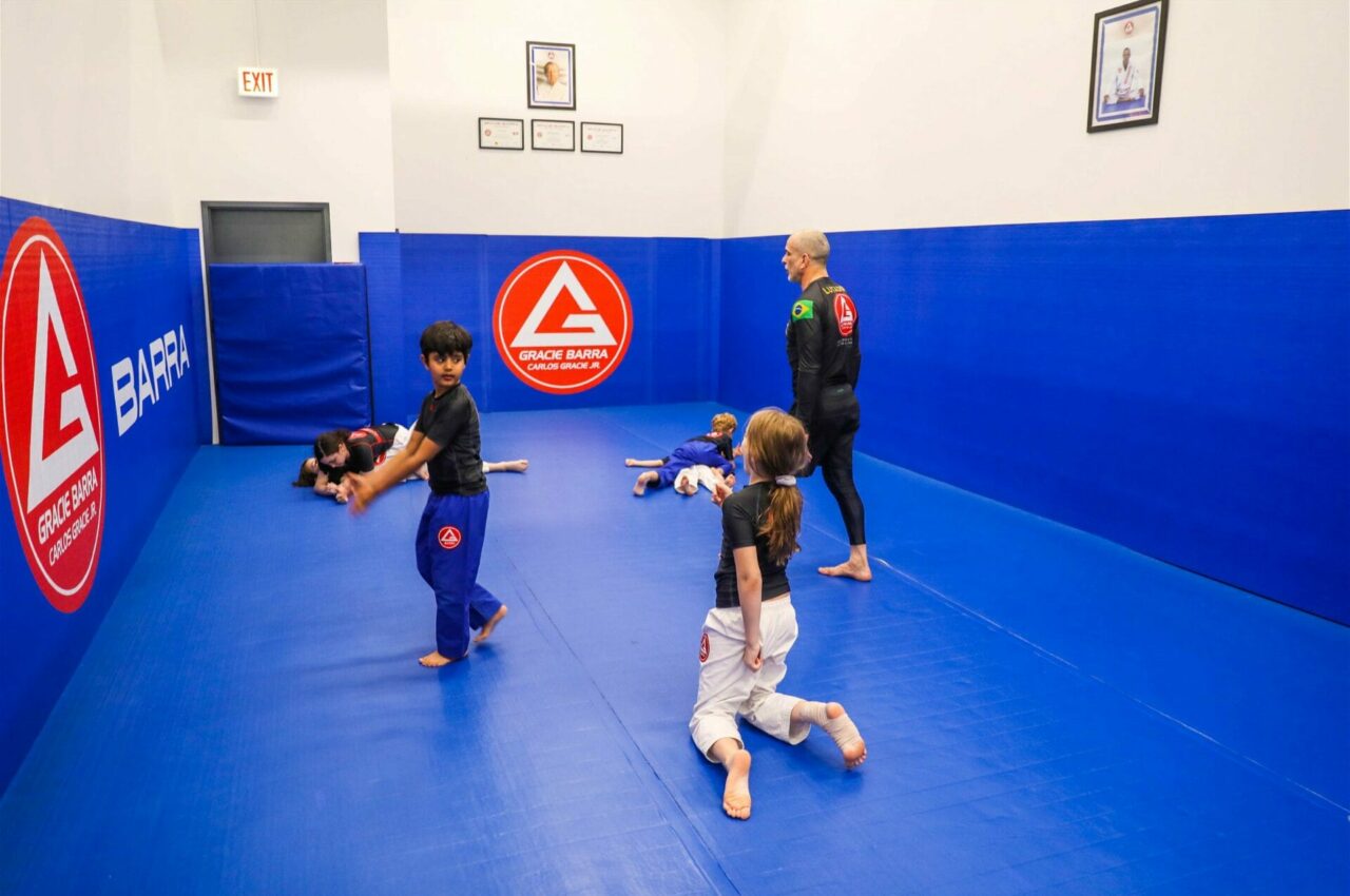 Children practice Brazilian Jiu-Jitsu and Muay Thai in a blue-matted room with an instructor, under the Gracie Barra logo on the wall.